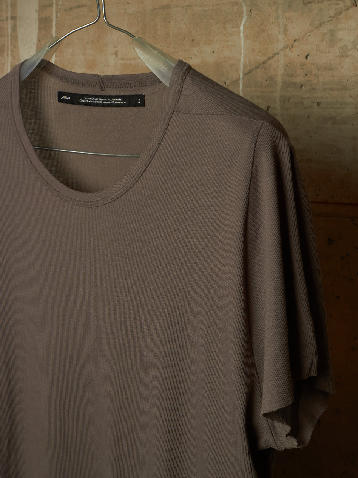 A taupe brown short-sleeve knit top hanging on a metal hanger against a textured stone wall background.