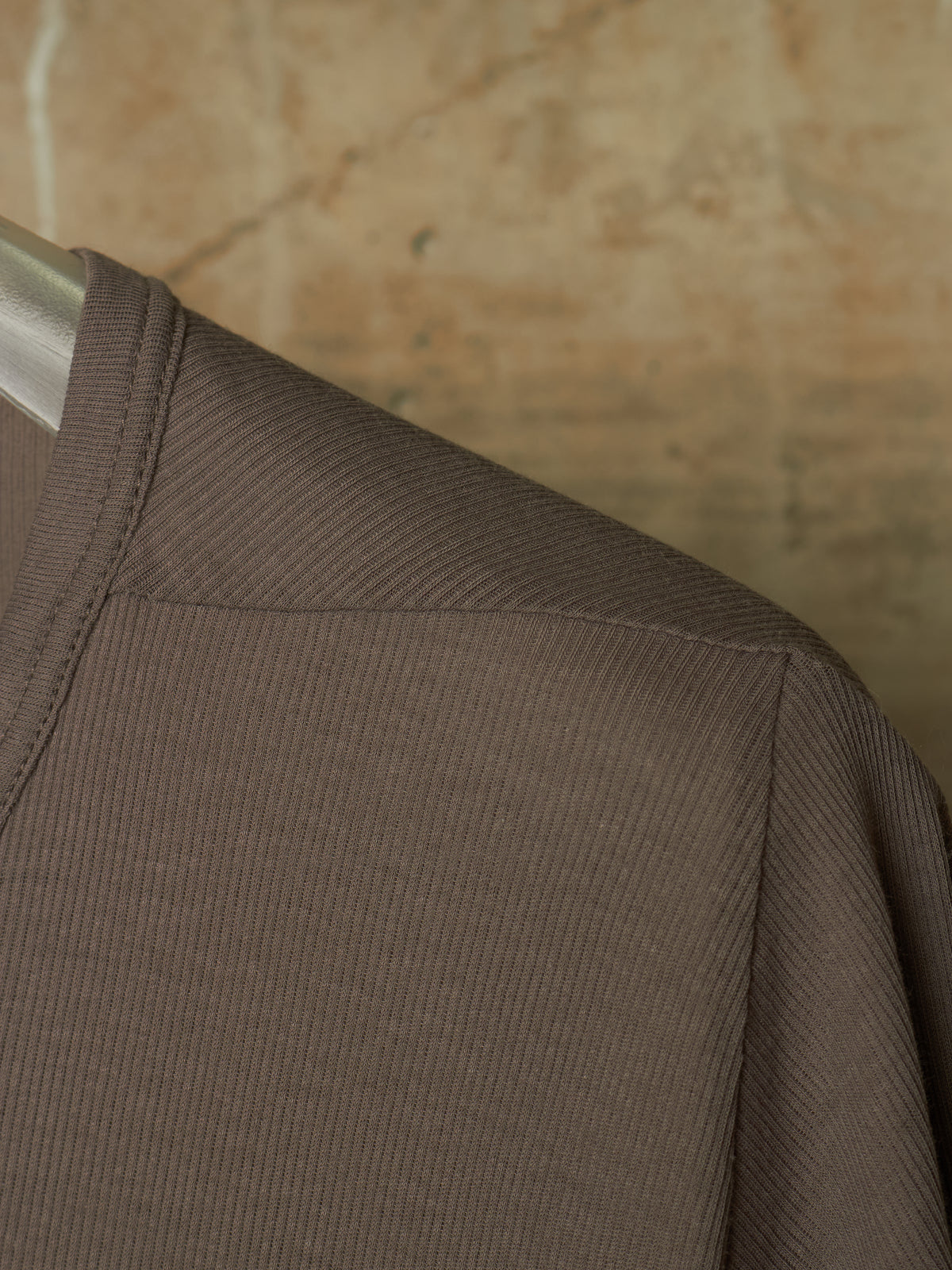 Close-up of a ribbed brown long-sleeve top on a hanger, showing the shoulder seam and textured fabric against a warm beige background.