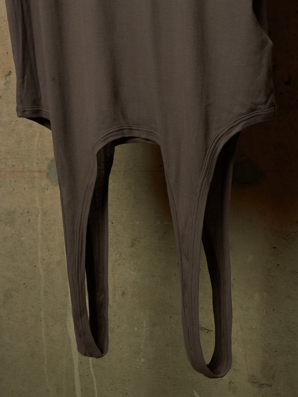 A dark brown ribbed bodysuit or long-sleeve top with split leg openings, hanging against a textured concrete wall.