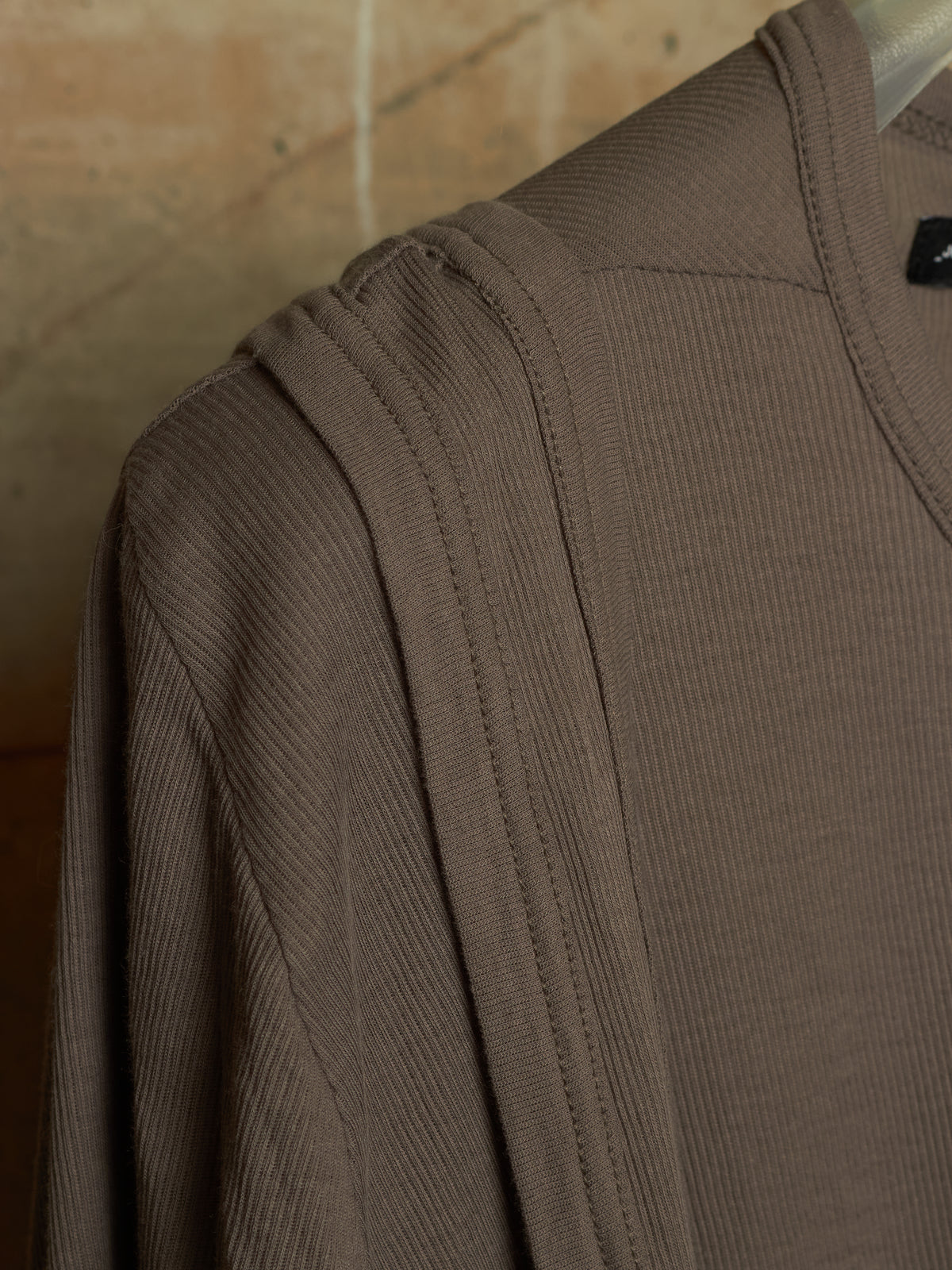 Close-up of a ribbed knit top in taupe brown, showing the shoulder seam and collar area against a warm beige background.