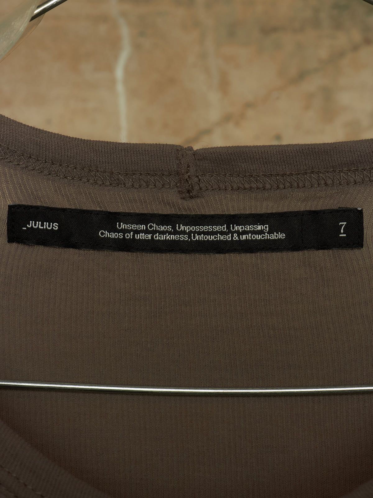 Close-up of a brown ribbed knit garment neck label reading JULIUS with white text and a size 7, shown on a hanger against a beige stone-like background.