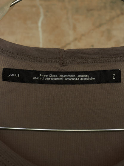 Close-up of a brown ribbed knit garment neck label reading JULIUS with white text and a size 7, shown on a hanger against a beige stone-like background.