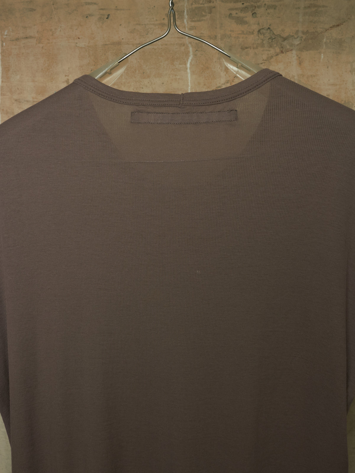 A dark brown long-sleeve knit top hanging on a clear plastic hanger against a textured beige wall.