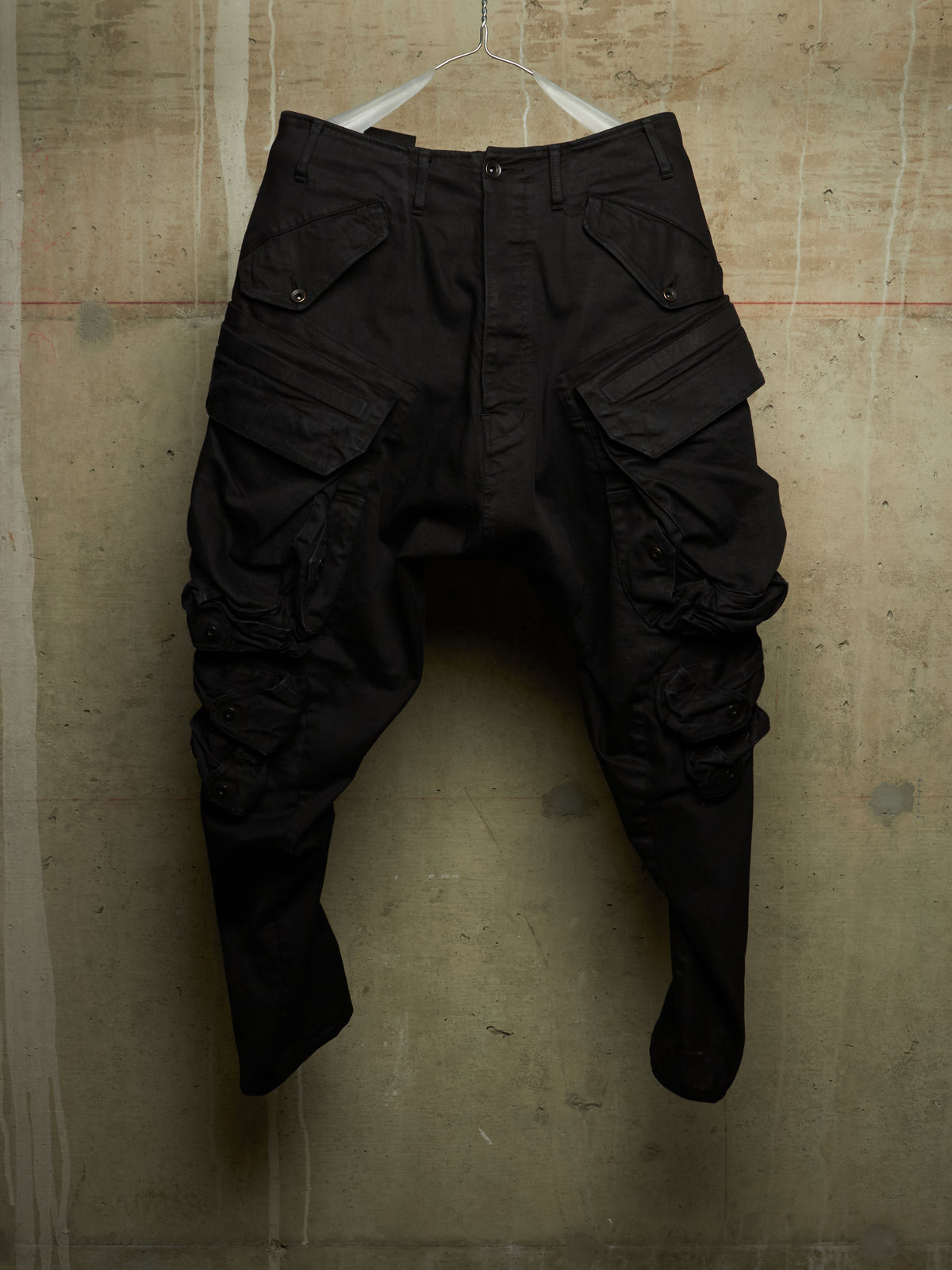 Black cargo pants with a unique crinkled, baggy fit, hanging on a white wire hanger against a textured concrete wall.
