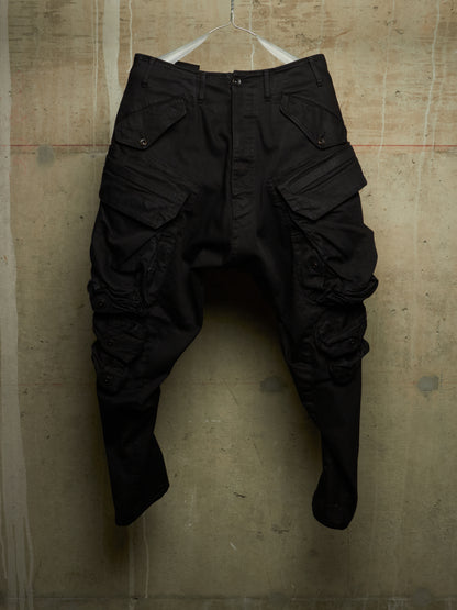 Black cargo pants with a unique crinkled, baggy fit, hanging on a white wire hanger against a textured concrete wall.
