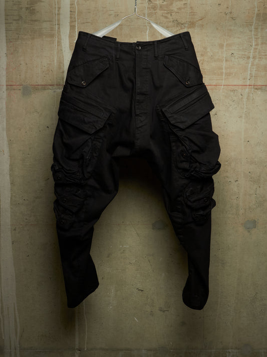 Black cargo pants with a unique crinkled, baggy fit, hanging on a white wire hanger against a textured concrete wall.