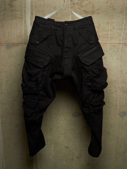 Black cargo pants with a unique sculptural, asymmetrical leg design, hanging on a wire hanger against a textured concrete wall.
