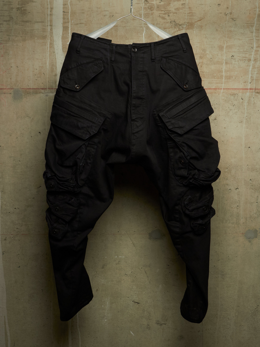 Black cargo pants with a unique sculptural, asymmetrical leg design, hanging on a wire hanger against a textured concrete wall.