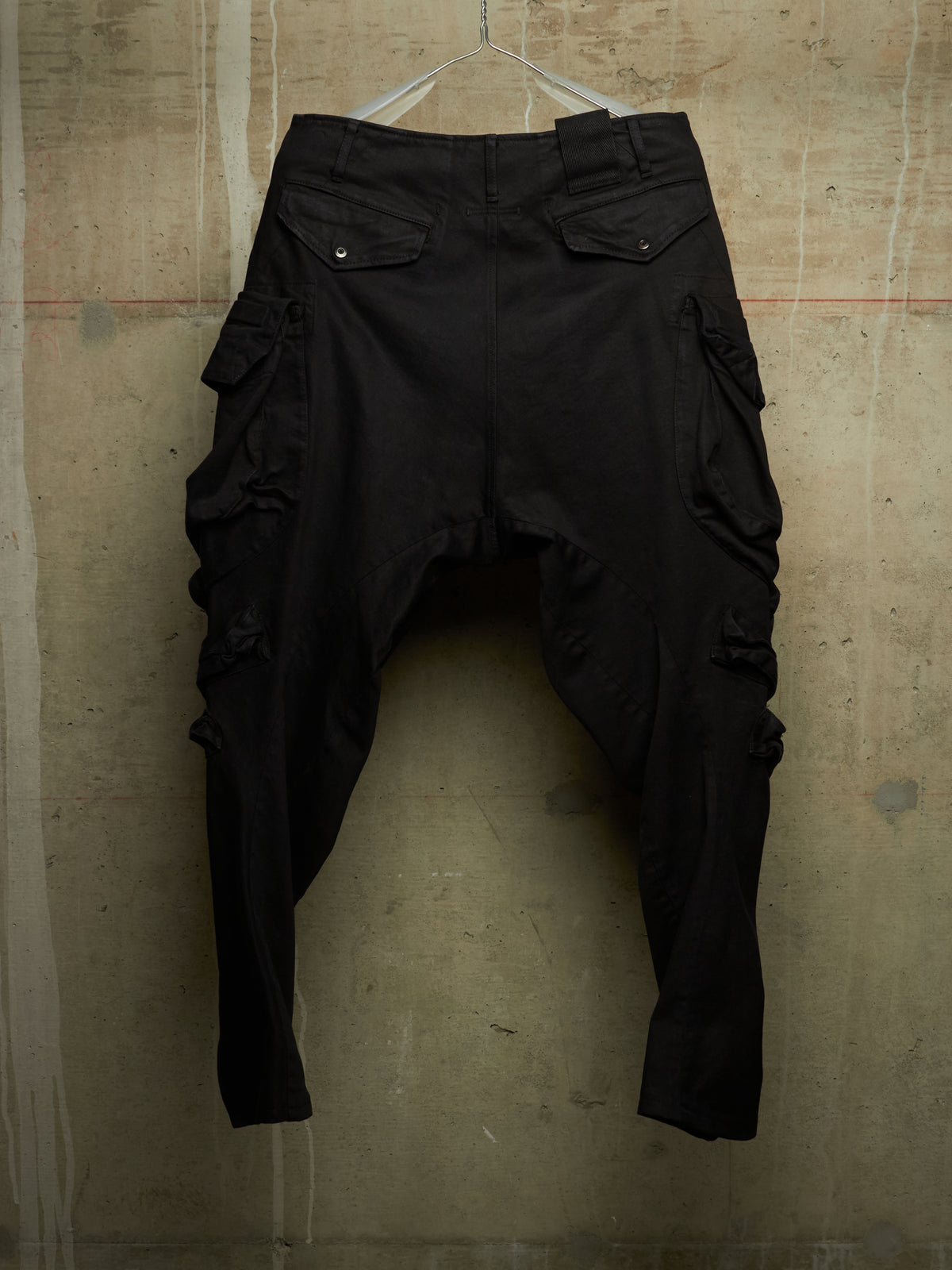 Black cargo-style pants with dramatic ruffled detailing on the legs, hanging against a textured concrete wall.