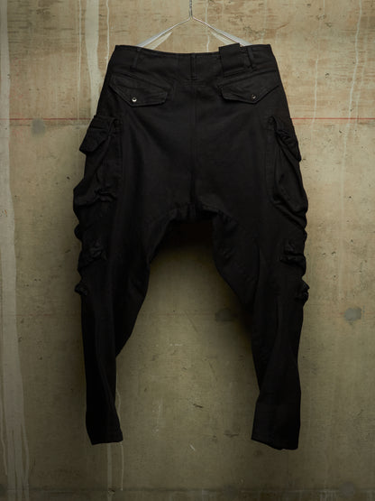 Black cargo-style pants with dramatic ruffled detailing on the legs, hanging against a textured concrete wall.