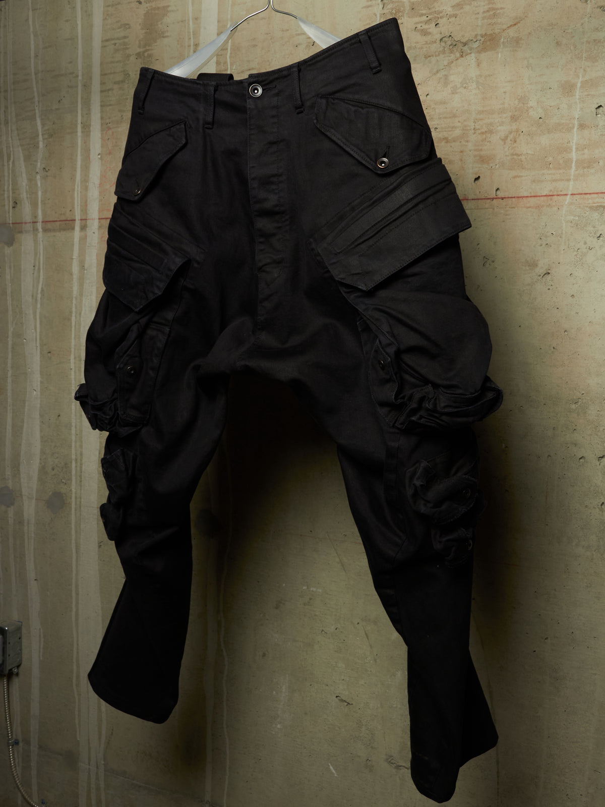 Black cargo pants with multiple flap pockets and a relaxed, baggy fit, hanging on a white hanger against a textured concrete wall.