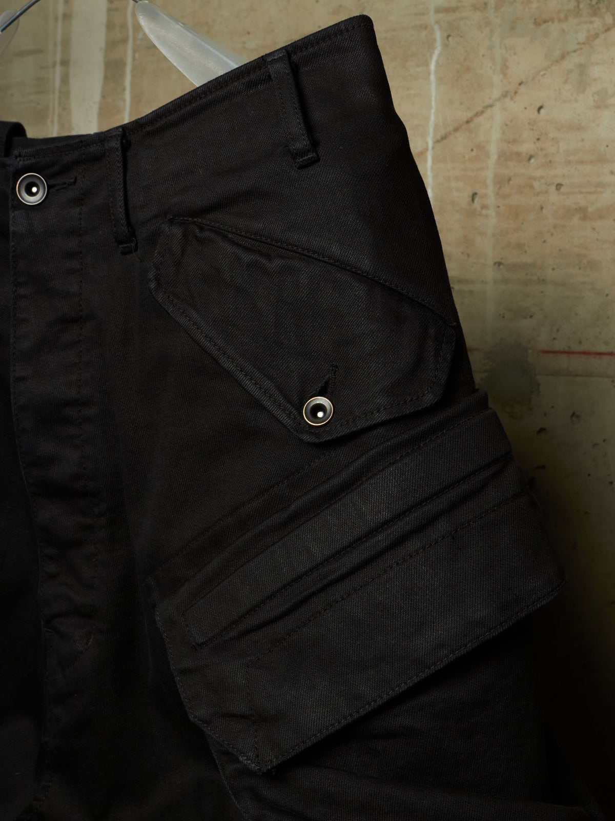 Close-up of black cargo pants showing a buttoned flap pocket and belt loops against a textured concrete background.