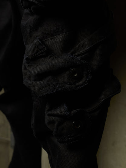 Close-up of black denim pants showing a rugged cargo pocket with raw frayed edges and metal snap closures.