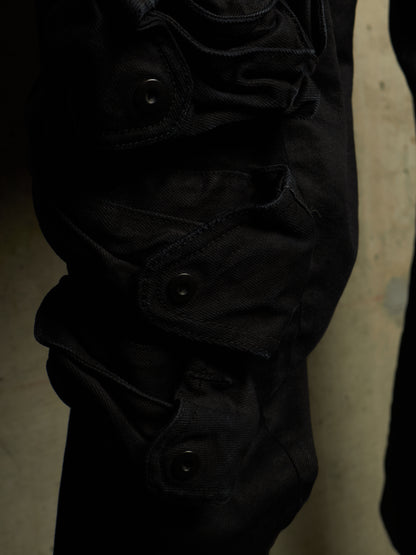 Close-up of black cargo pants showing the side pocket details with snap buttons, set against a dark, moody background.