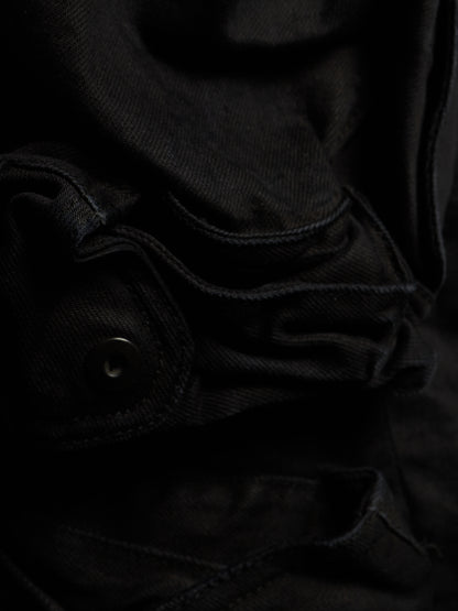 Close-up detail of a black textured fabric garment, showing a buttoned flap pocket and layered seams.