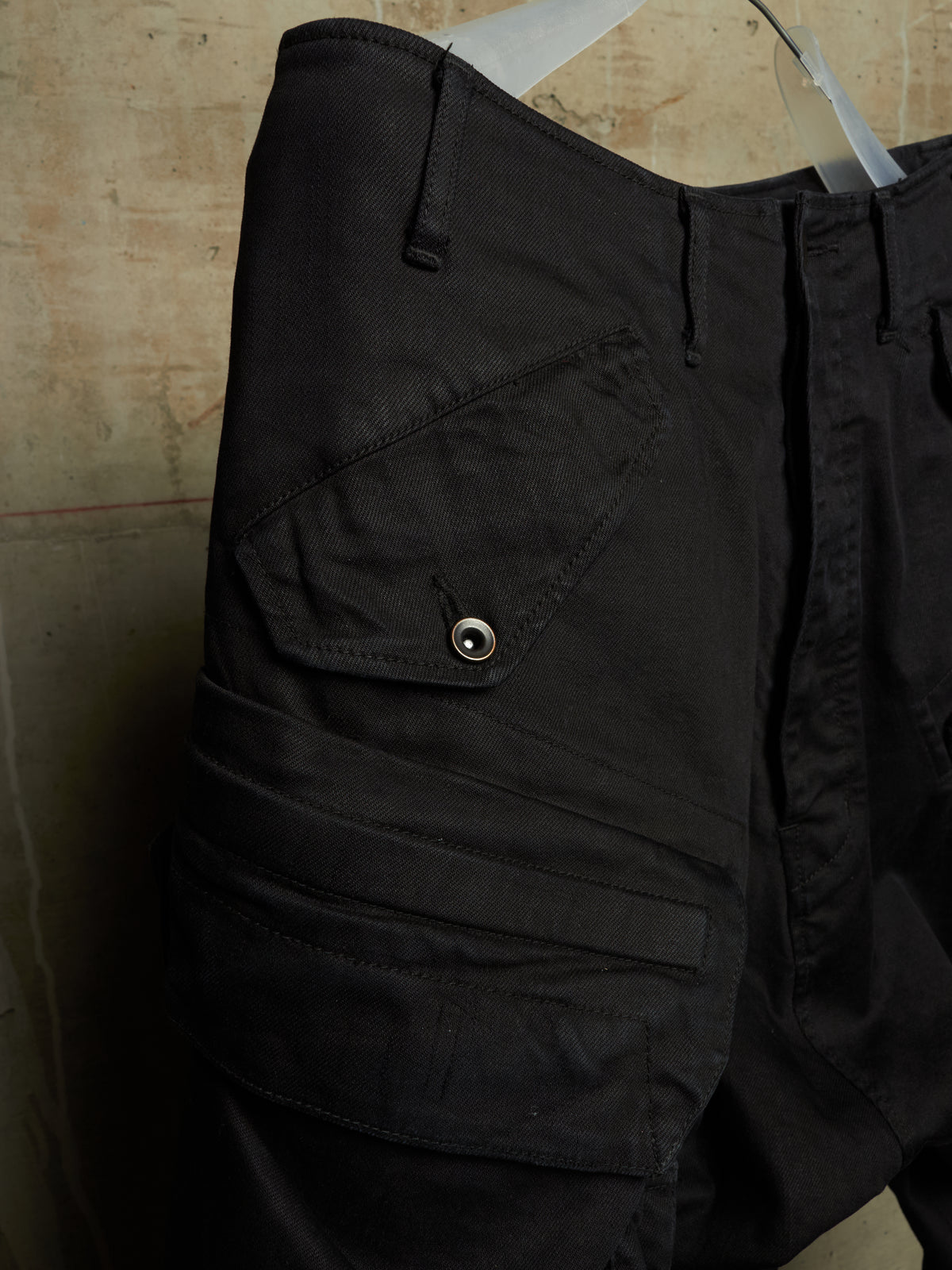 Close-up of black cargo shorts with multiple flap pockets and a metal snap button, hanging against a textured beige wall.