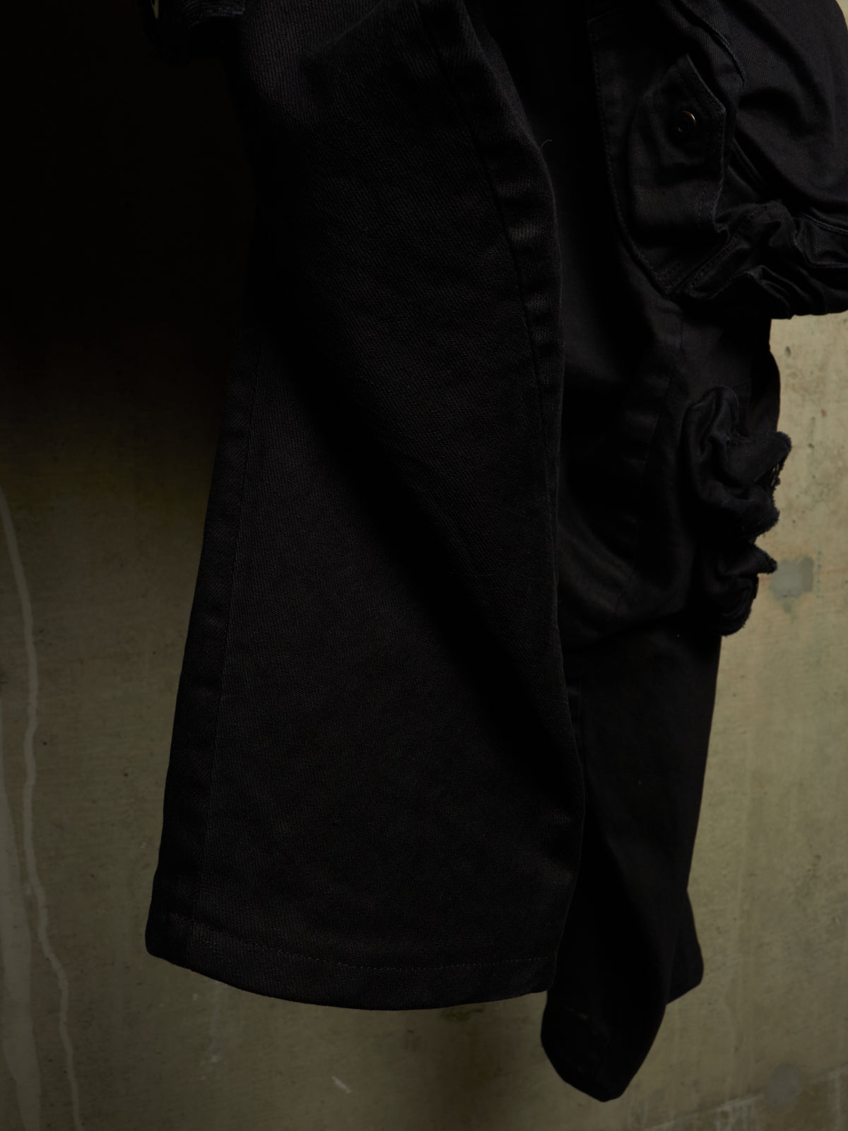 A close-up of a black garment, likely a jacket or coat, featuring a large ruffled or gathered detail on the right side, shown against a textured concrete wall.