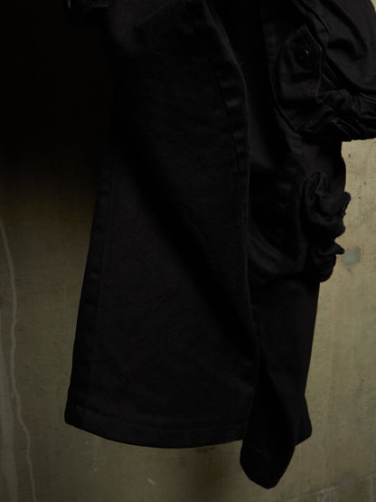 A close-up of a black garment, likely a jacket or coat, featuring a large ruffled or gathered detail on the right side, shown against a textured concrete wall.