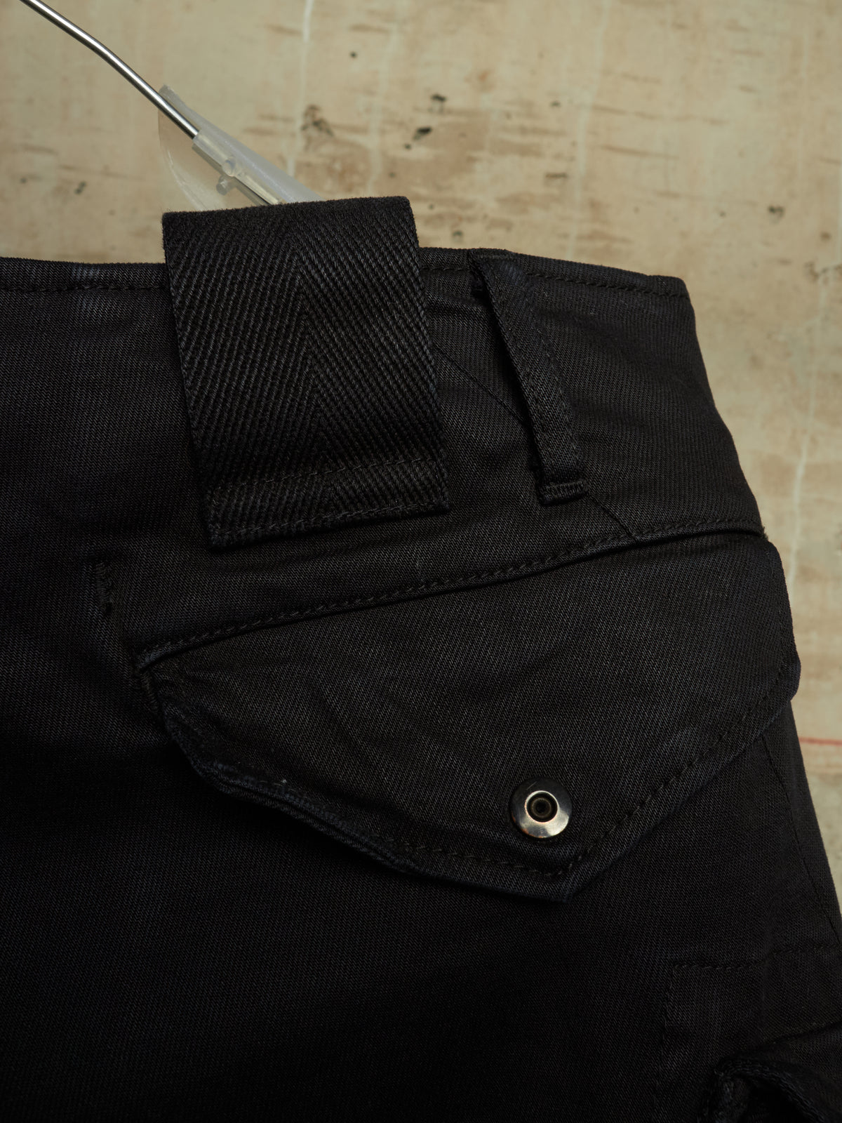 Close-up of black denim pants showing the waistband, belt loop, and a flap pocket with a metal snap button, set against a light textured background.