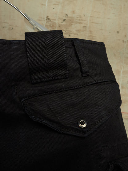 Close-up of black denim pants showing the waistband, belt loop, and a flap pocket with a metal snap button, set against a light textured background.