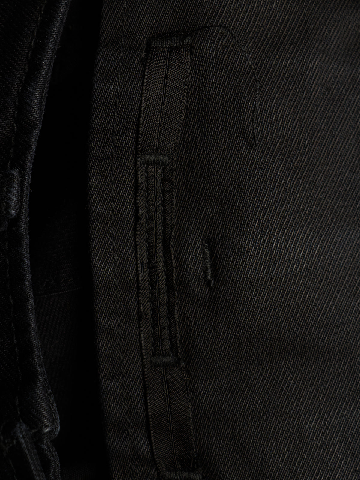 Close-up detail of a black textured fabric garment, showing a vertical stitched strap or zipper pull on a dark woven textile.