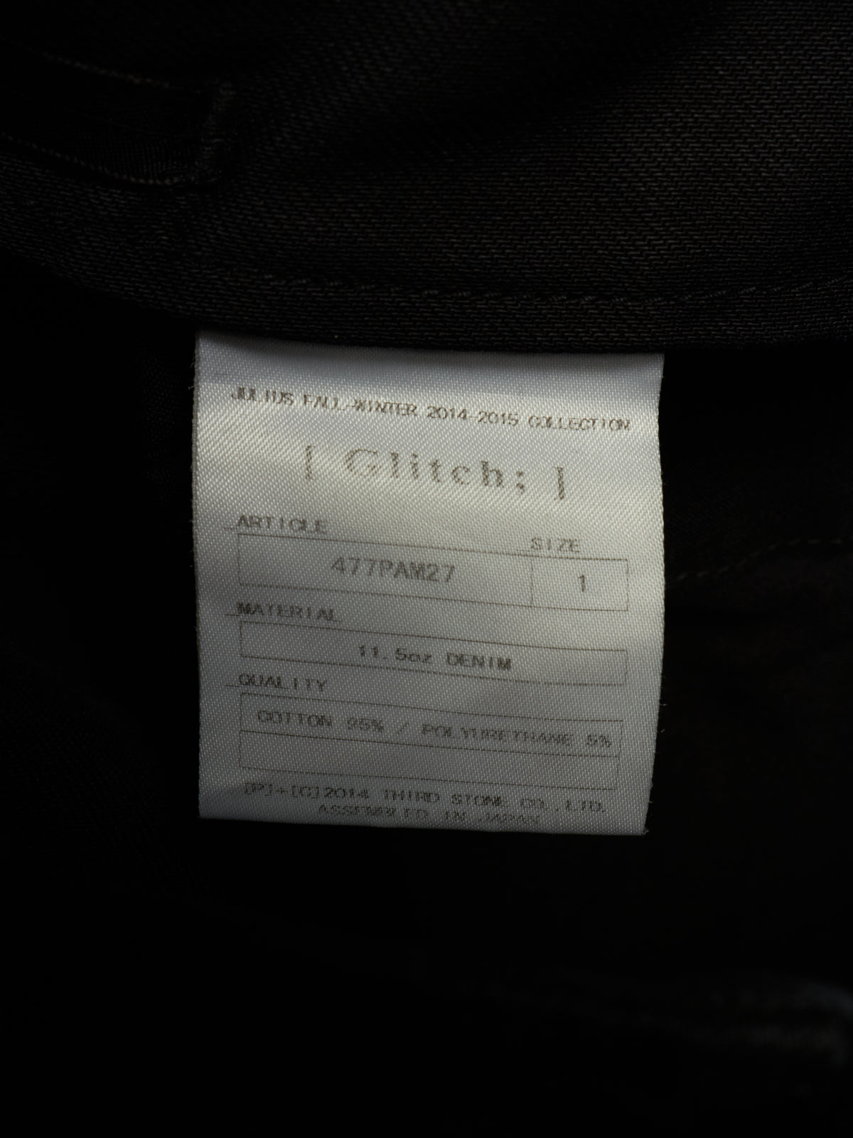 A close-up of a white fabric product label inside a black garment, showing the brand name 'Glitch;' and product details.