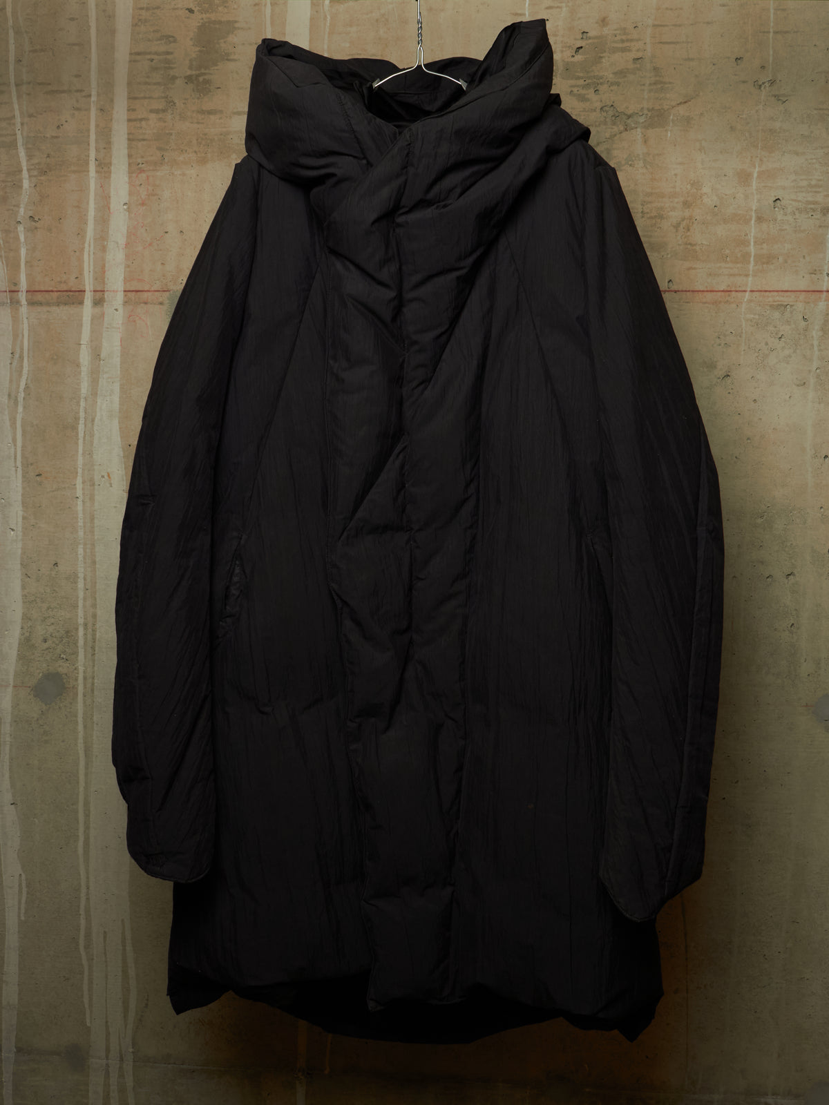 A black oversized quilted puffer jacket with a high draped collar, hanging against a textured concrete wall.