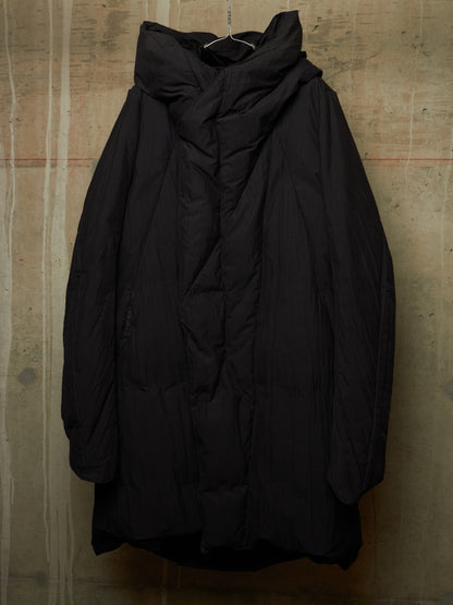 A black oversized quilted puffer jacket with a high draped collar, hanging against a textured concrete wall.