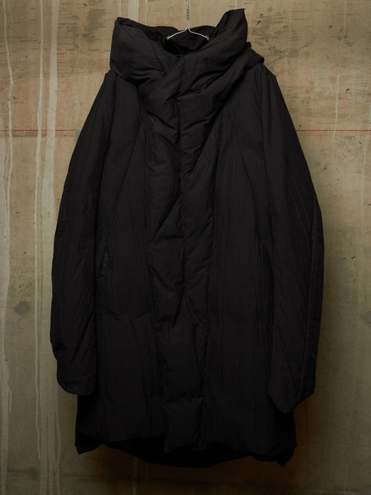 A black oversized quilted puffer jacket with a high draped collar, hanging against a textured concrete wall.