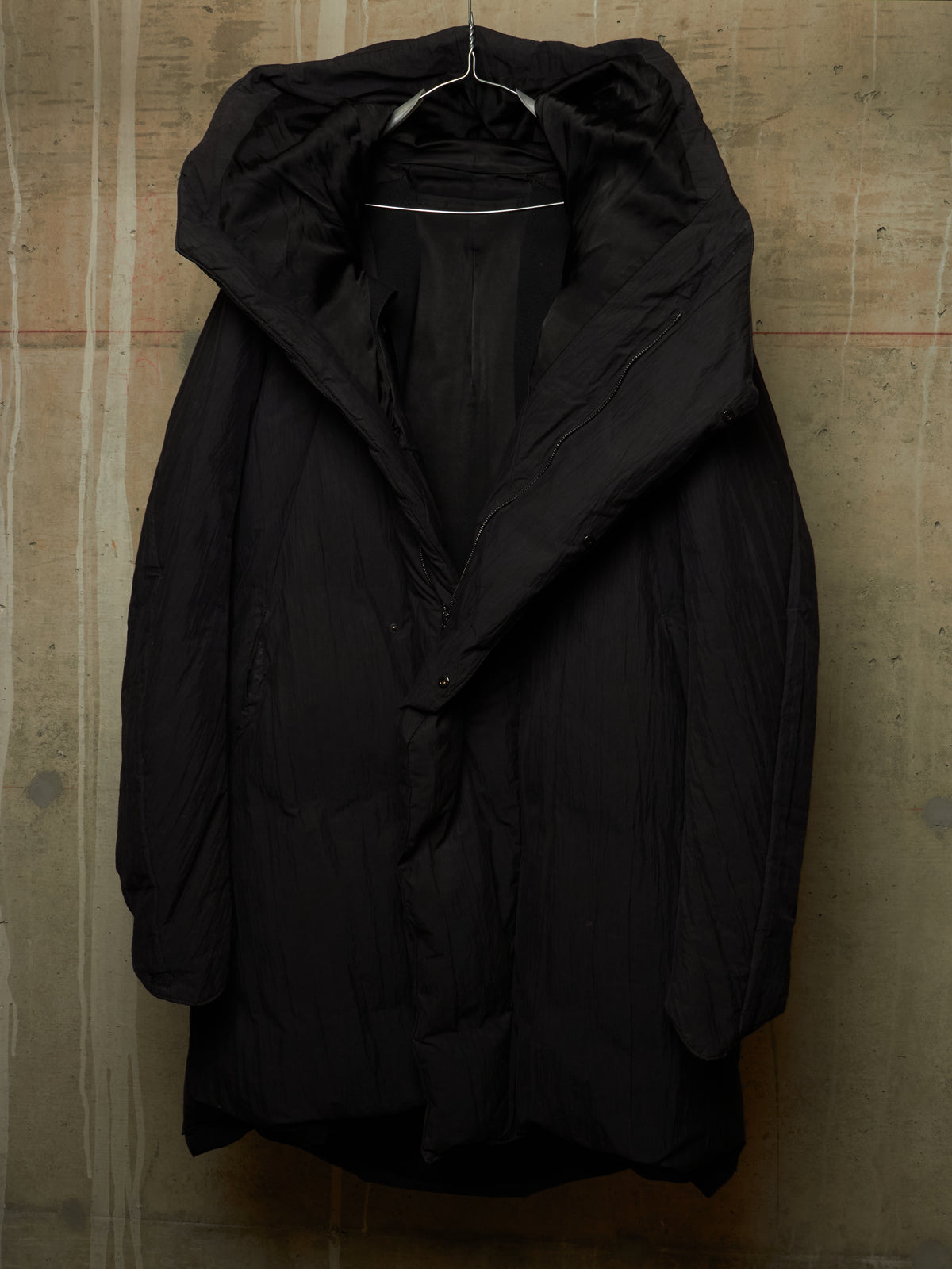 A black hooded puffer jacket hanging on a metal hanger against a textured concrete wall.