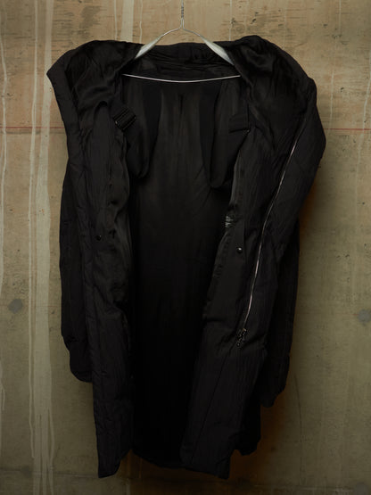 A black hooded puffer jacket with a high collar and front zipper, hanging on a metal hanger against a textured concrete wall.