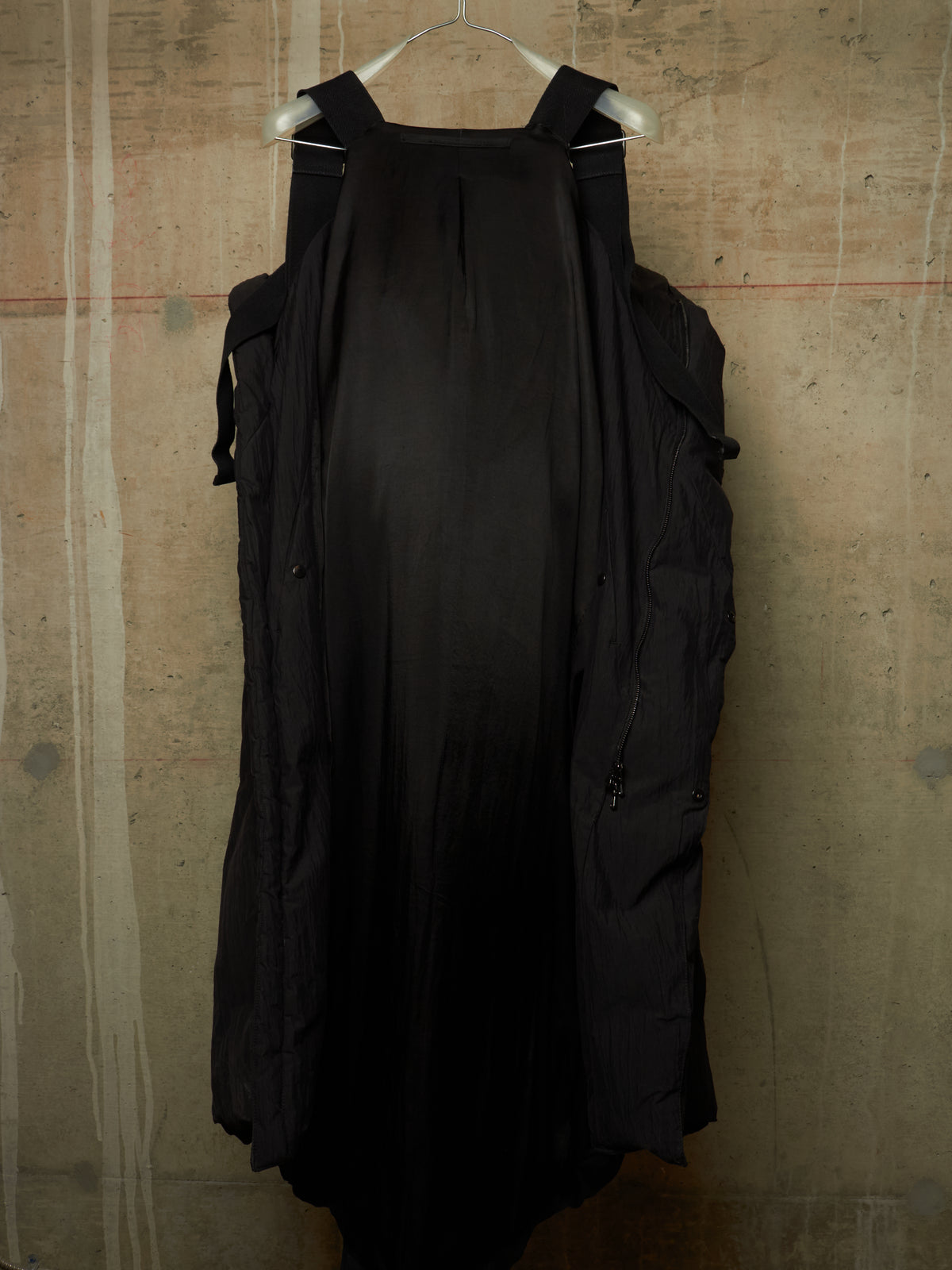 A black sleeveless dress with shoulder straps and side pockets, hanging on a white hanger against a textured concrete wall.