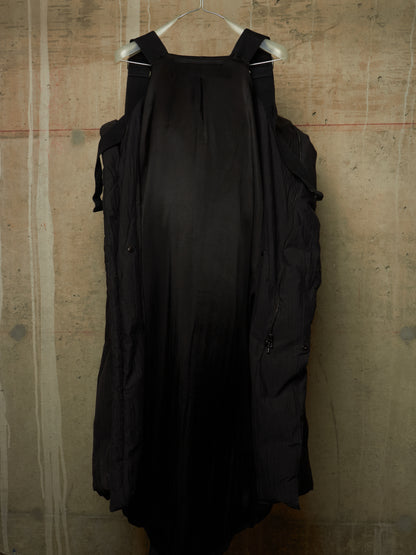 A black sleeveless dress with shoulder straps and side pockets, hanging on a white hanger against a textured concrete wall.