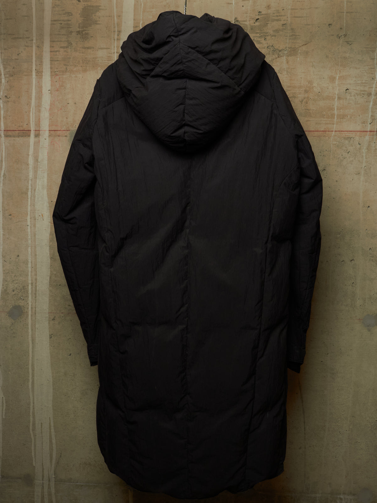A long black hooded puffer jacket shown from the back, hanging against a textured concrete wall.
