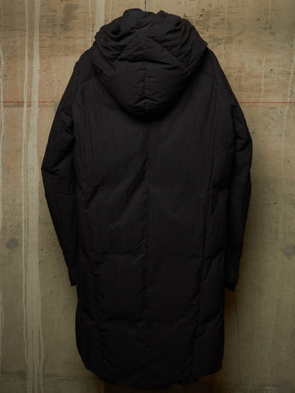 A long black hooded puffer jacket shown from the back, hanging against a textured concrete wall.