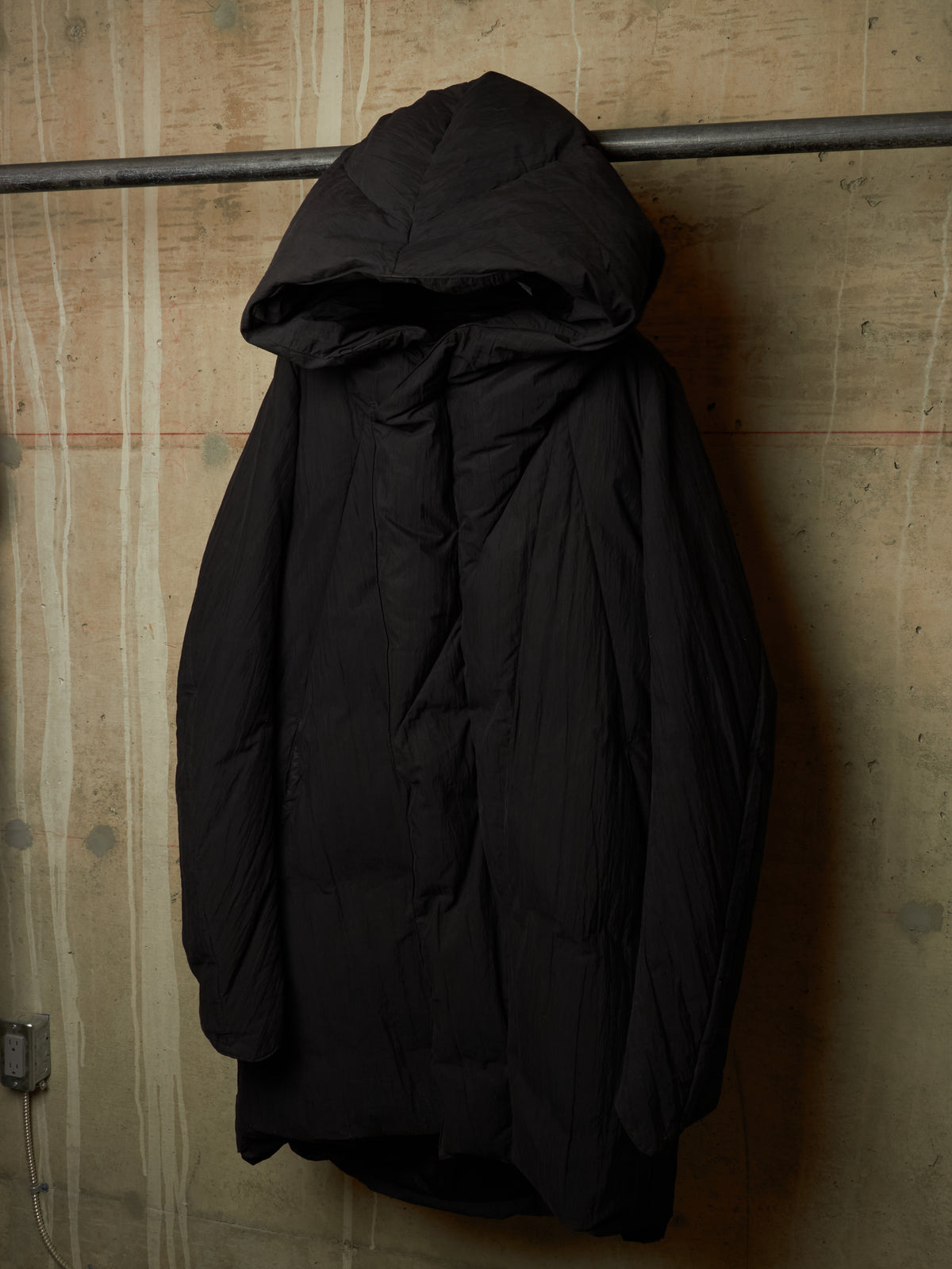 A black hooded puffer jacket hanging on a metal rack against a textured concrete wall.
