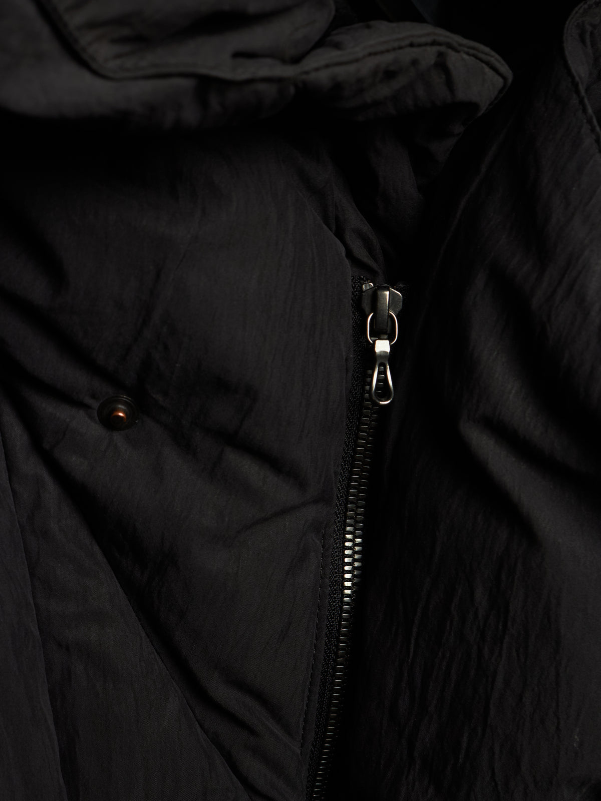 Close-up of a black puffer jacket showing the textured crinkled fabric, a silver metal zipper, and a small dark button detail.