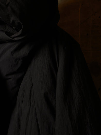 A close-up of a black hooded jacket or coat with a crinkled texture, shown in very dark lighting against a dim brown background.