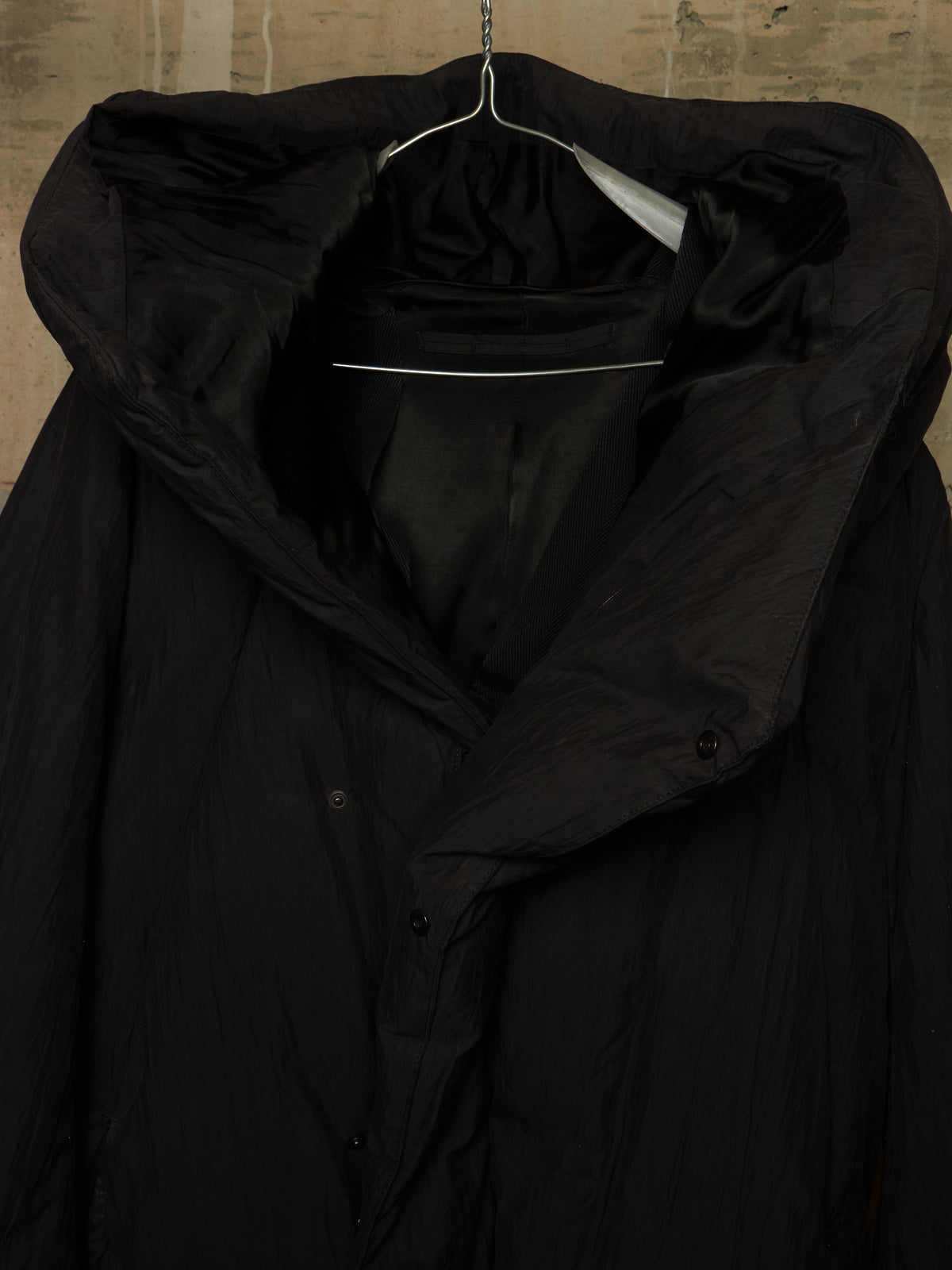 A black hooded puffer jacket with a high collar and button front, hanging on a metal hanger against a textured beige wall.