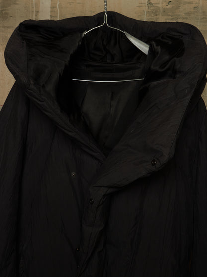 A black hooded puffer jacket with a high collar and button front, hanging on a metal hanger against a textured beige wall.