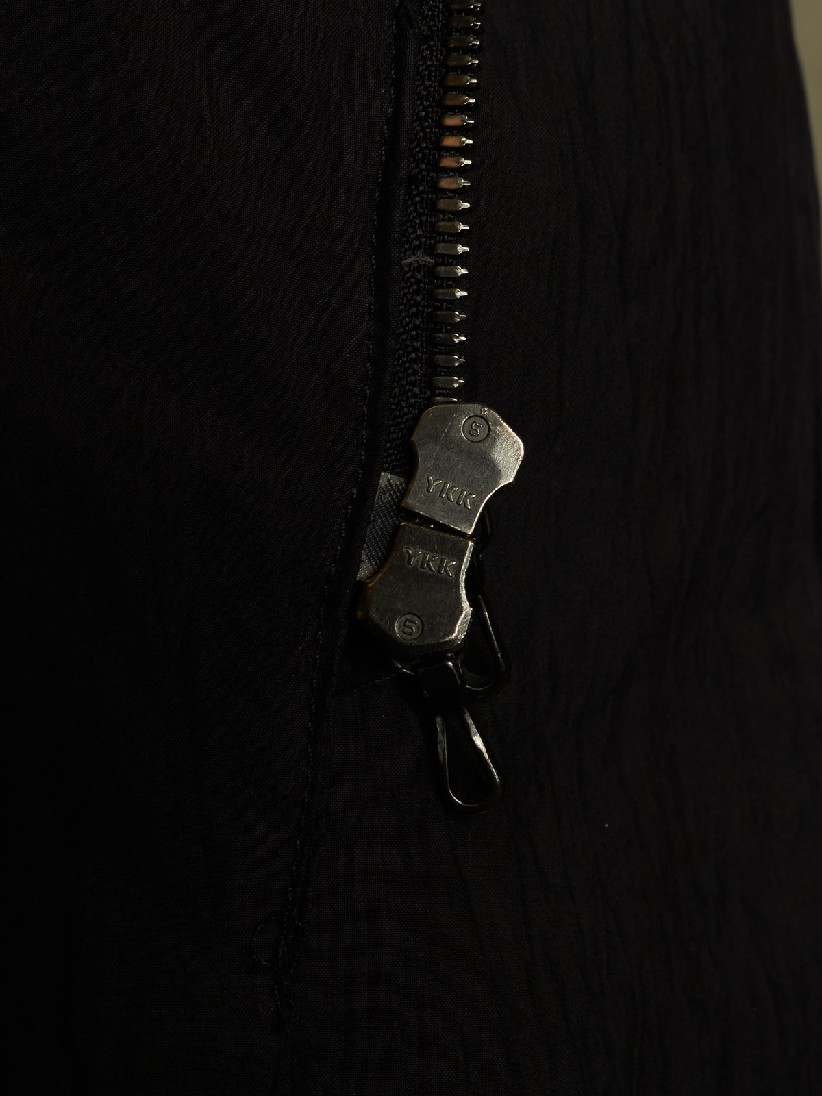 Close-up of a black zippered garment with a dark metal zipper pull and a small attached black clip, shown against a very dark background.