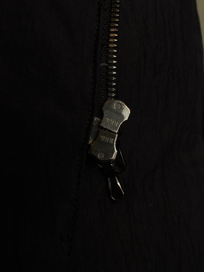 Close-up of a black zippered garment with a dark metal zipper pull and a small attached black clip, shown against a very dark background.