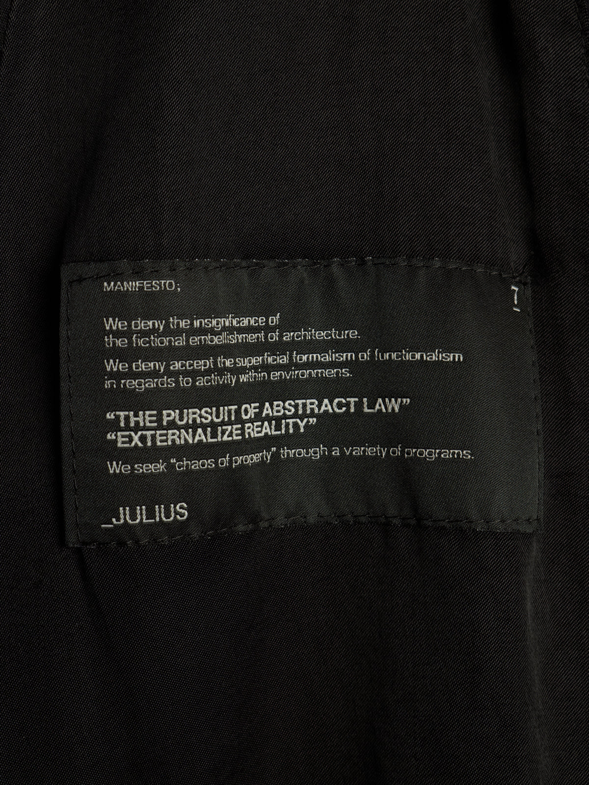 Close-up of a black garment label with white printed text reading 'MANIFESTO' and 'JULIUS' on a dark fabric background.