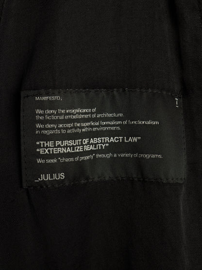 Close-up of a black garment label with white printed text reading 'MANIFESTO' and 'JULIUS' on a dark fabric background.