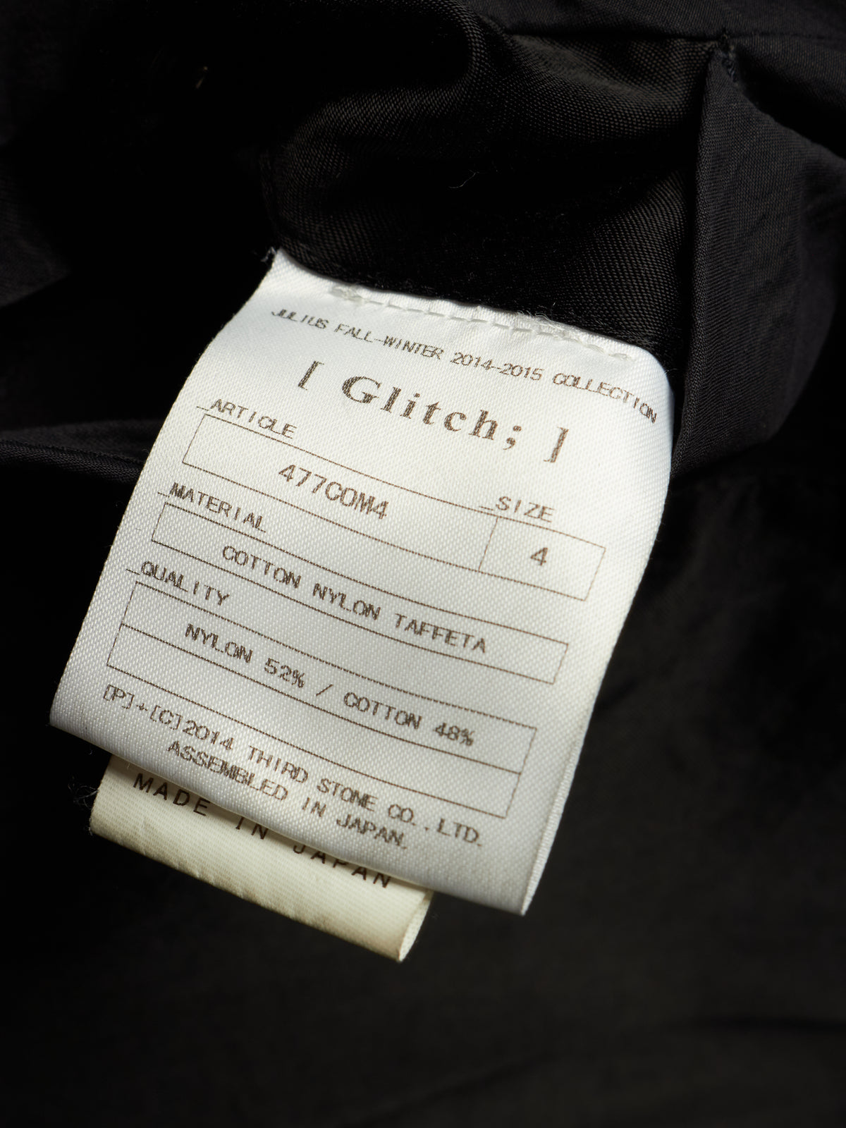 A close-up of a white fabric garment label sewn onto a black garment, showing text including '[ Glitch; ],' material details, and 'MADE IN JAPAN.'.