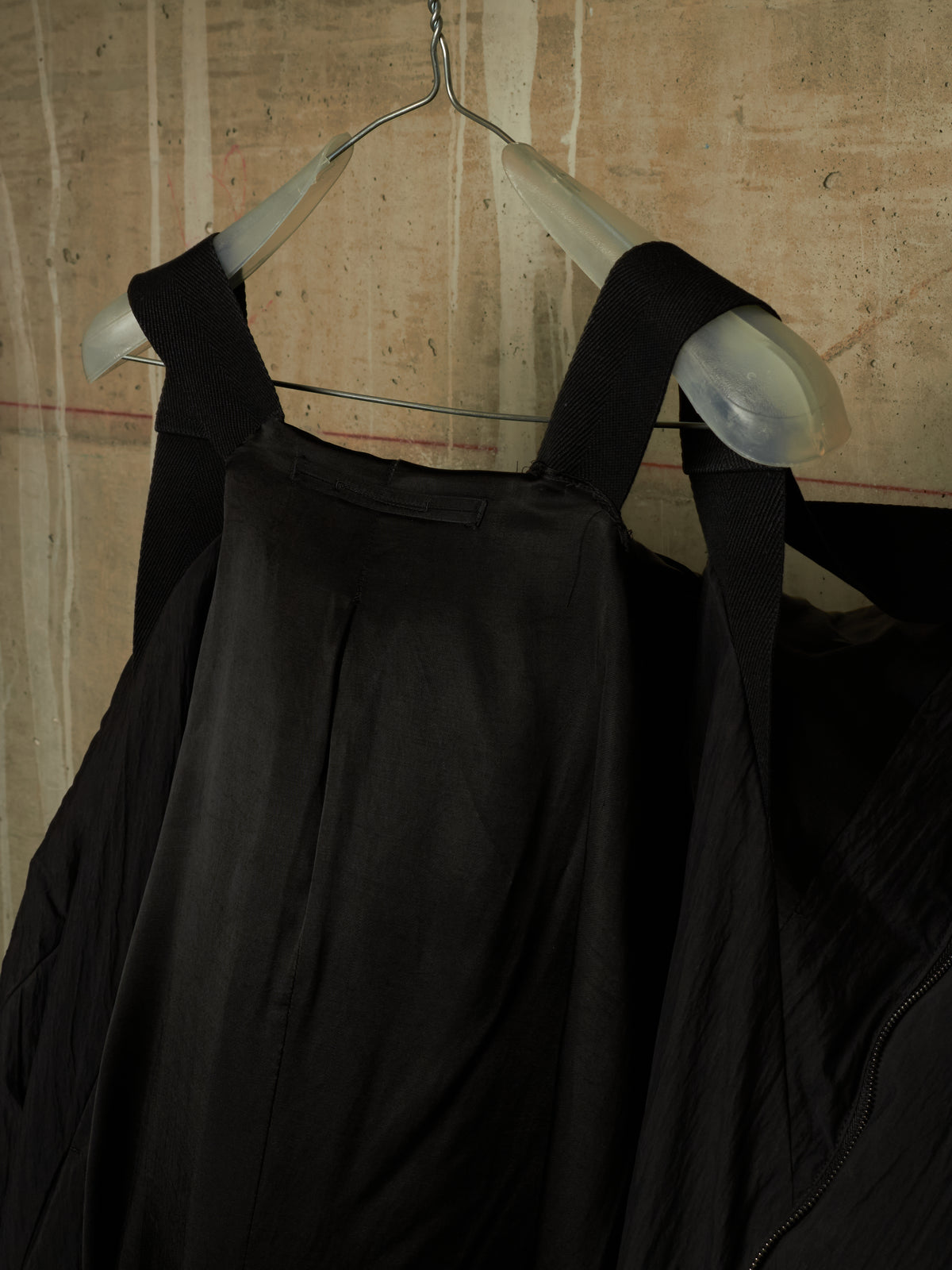 A black sleeveless garment with wide shoulder straps hanging on a clear plastic hanger against a textured beige wall.