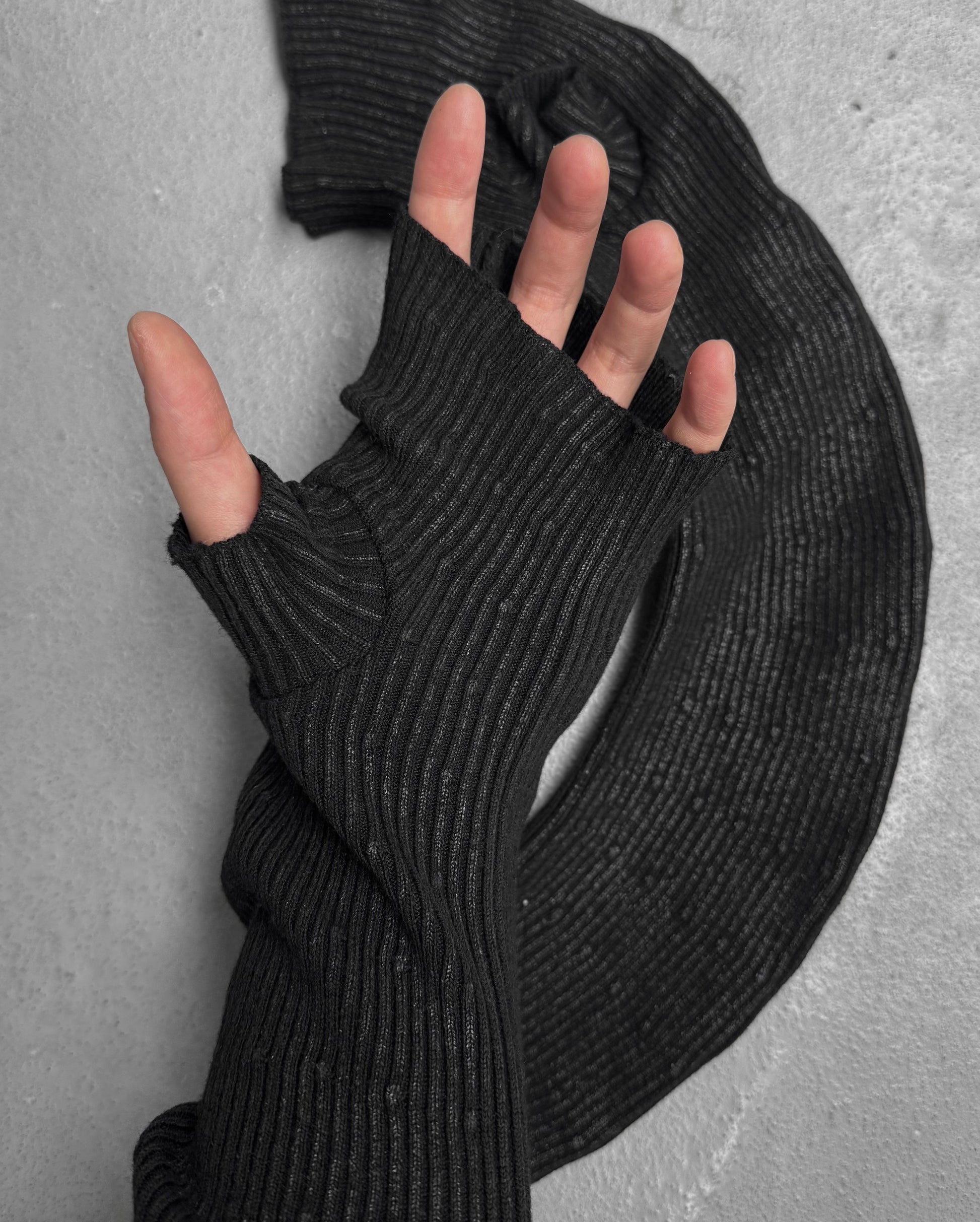 A hand wearing DRIEW’s Julius_7 AW24-25 “rift;” Curved Wax Coated black, ribbed, fingerless glove with a thumb hole rests on a textured gray surface, matching the fabric coiled beside it. One size; extends up the arm.