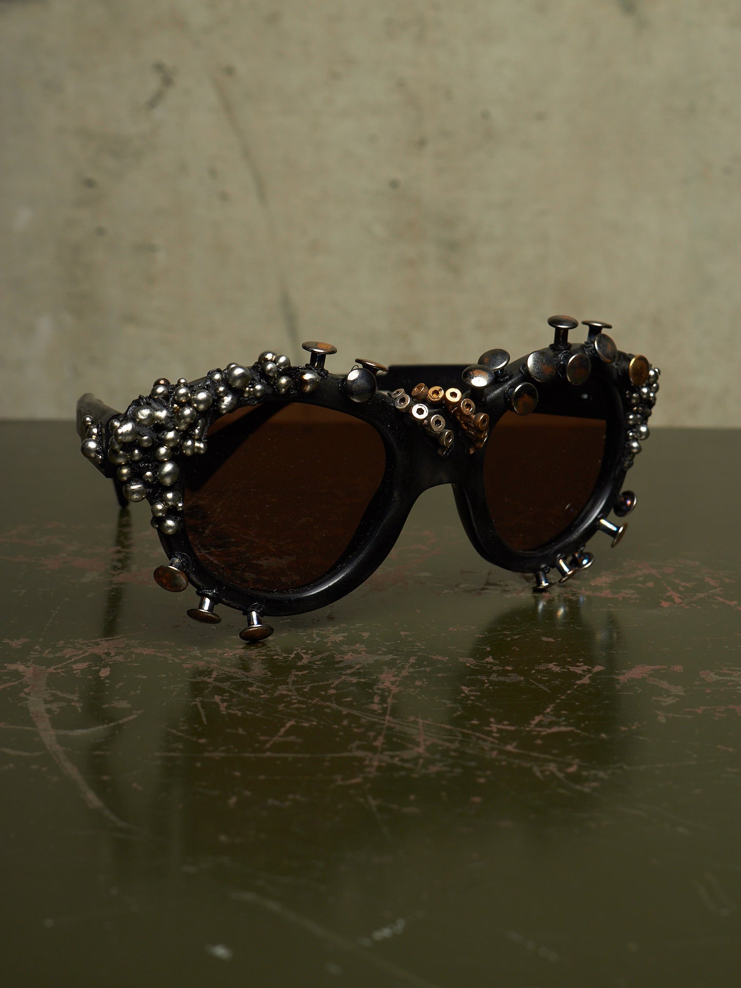 Studded gothic sunglasses with dark brown lenses and silver-toned metal studs and beads on a dark reflective surface.