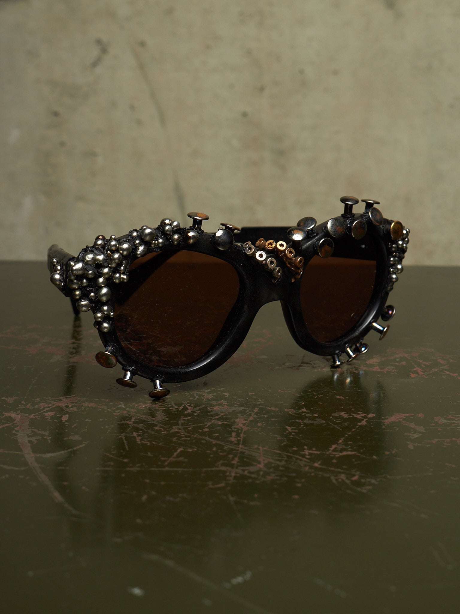 Studded gothic sunglasses with dark brown lenses and silver-toned metal studs and beads on a dark reflective surface.