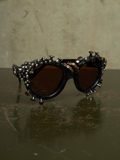 Studded gothic sunglasses with dark brown lenses and silver-toned metal studs and beads on a dark reflective surface.